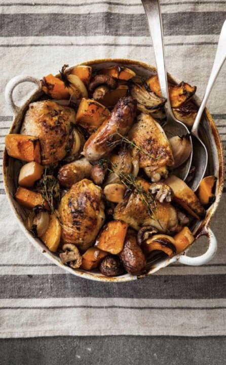 A rustic chicken and squash traybake inspired by British classics, with roast chicken, sausages, pumpkin, onions, and mushrooms garnished with herbs in a white round casserole dish on a striped cloth background.