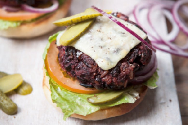 A Hairy Dieters Veggie Burger with a patty, melted cheese, lettuce, tomato slice, pickle slices, and red onion on a bun, partially assembled on a wooden surface.
