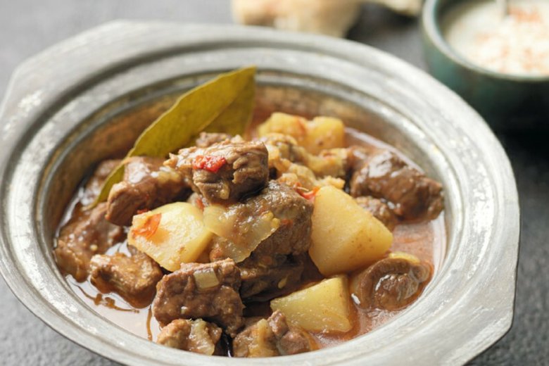 A bowl of Lamb Vindaloo with potatoes and bread on a table from the Hairy Bikers' Great Curries recipe book