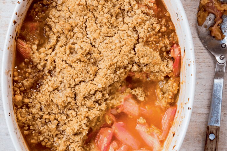 An oval baking dish containing a rhubarb and orange crumble with visible rhubarb pieces and golden brown topping, alongside a used serving spoon.