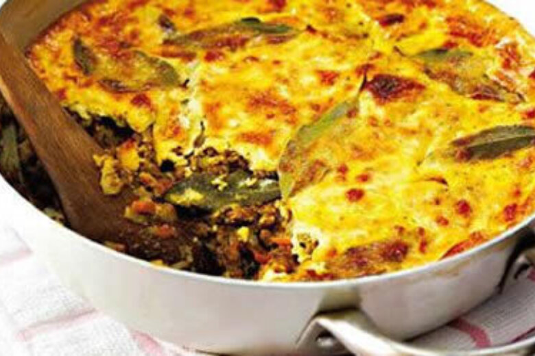 A golden brown baked dish, partially served with a wooden spoon, reveals layers of ingredients. This Beef Mince Bobotie, inspired by the Hairy Bikers Cookbook, includes a mix of vegetables and meat, all elegantly topped with bay leaves.