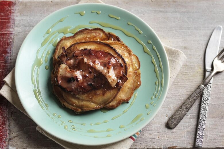 A stack of banana & bacon pancakes topped with grilled bacon and drizzled with syrup on a light green plate next to a fork and knife on a light wooden table, reminiscent of the Hairy Bikers' homey style.