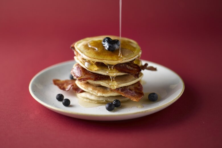 A stack of banana & bacon pancakes topped with grilled bacon and drizzled with syrup on a light green plate next to a fork and knife on a light wooden table, reminiscent of the Hairy Bikers' homey style.