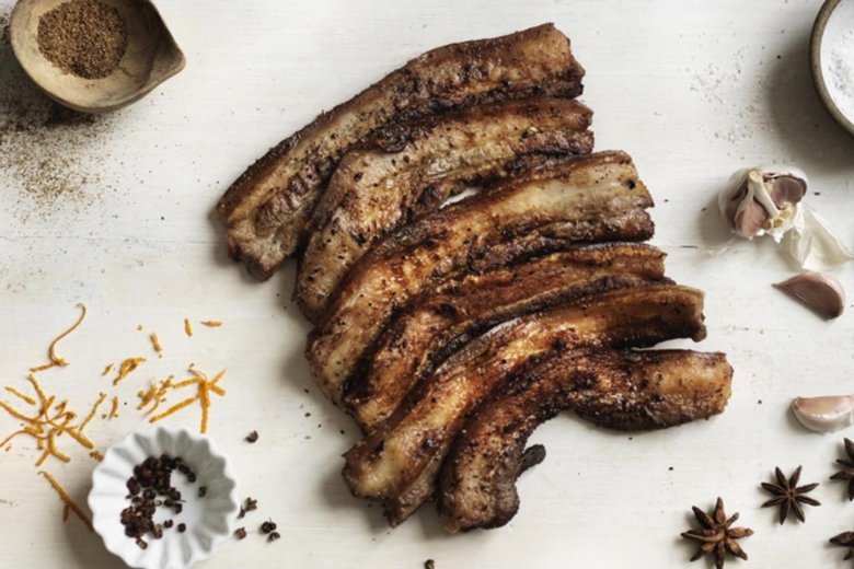 Barbecued belly pork on a table surrounded by spices, garlic, and pieces of orange zest, inspired by the Best of British cookery book from The Hairy Bikers