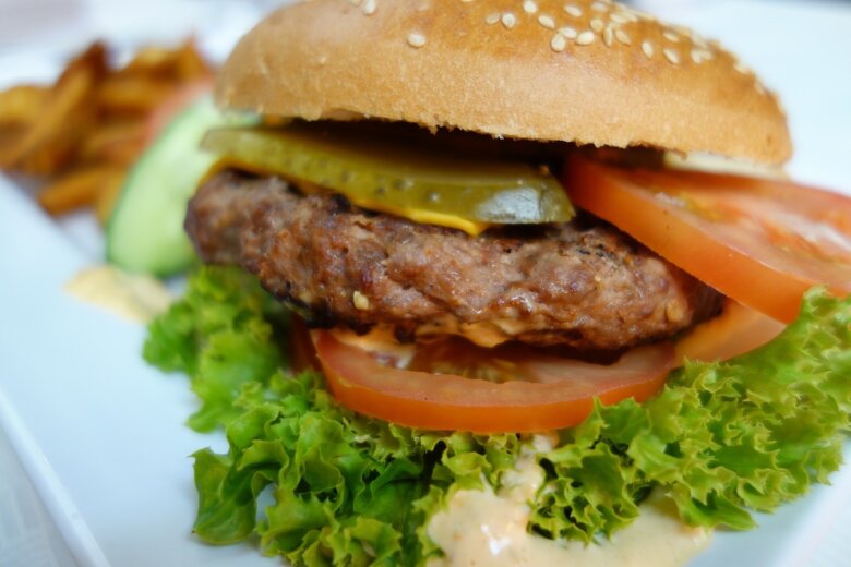 A Biker burger featuring a beef patty with lettuce, tomato, pickles,  in a sesame seed bun, served on a white plate with fries in the background. This mouthwatering delight is straight from the Hairy Bikers Recipe collection.