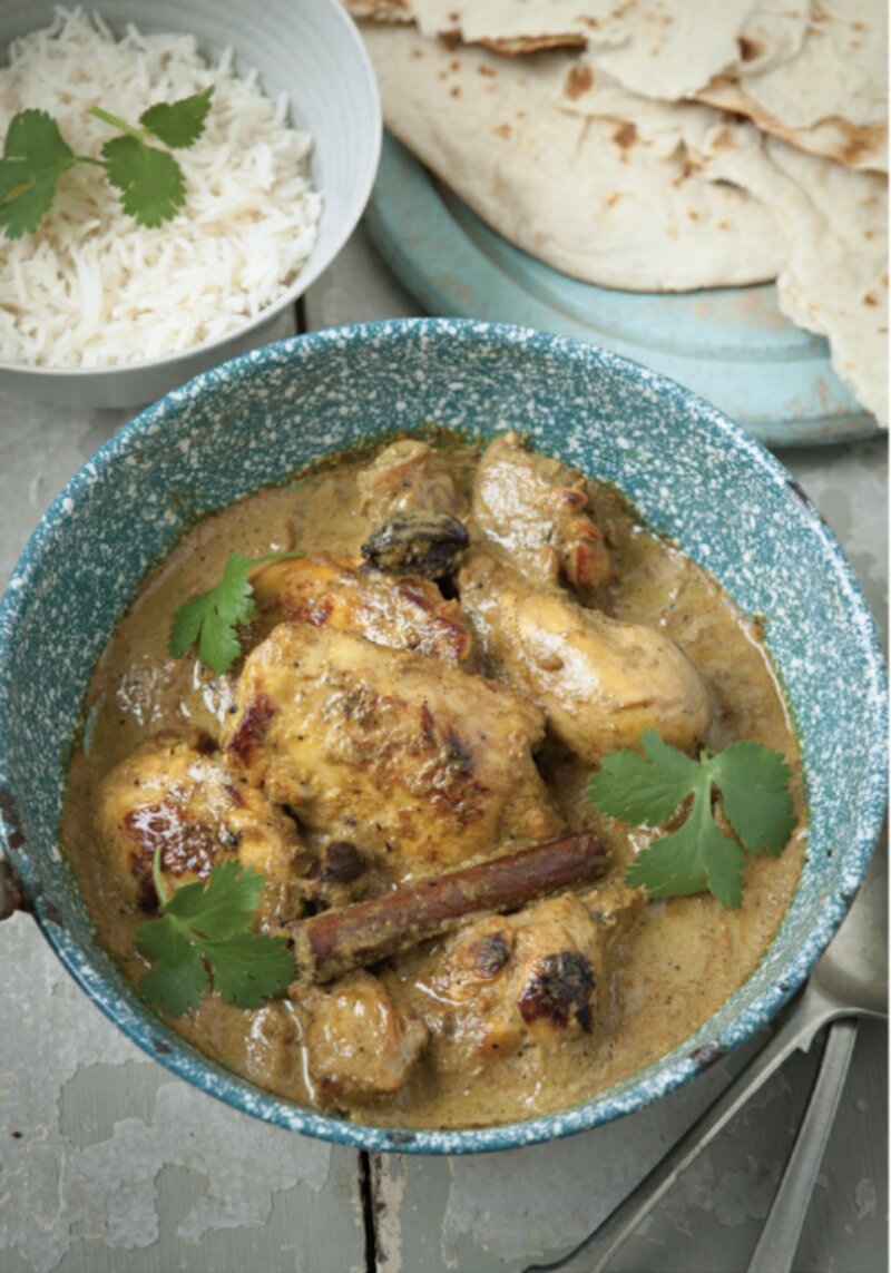 A blue bowl filled with Shorshe Murgi, a curry garnished with coriander, accompanied by a side of white rice and flatbread in the background—a dish perfect for fans of 'Chicken & Egg cookbook by the Hairy Bikers.