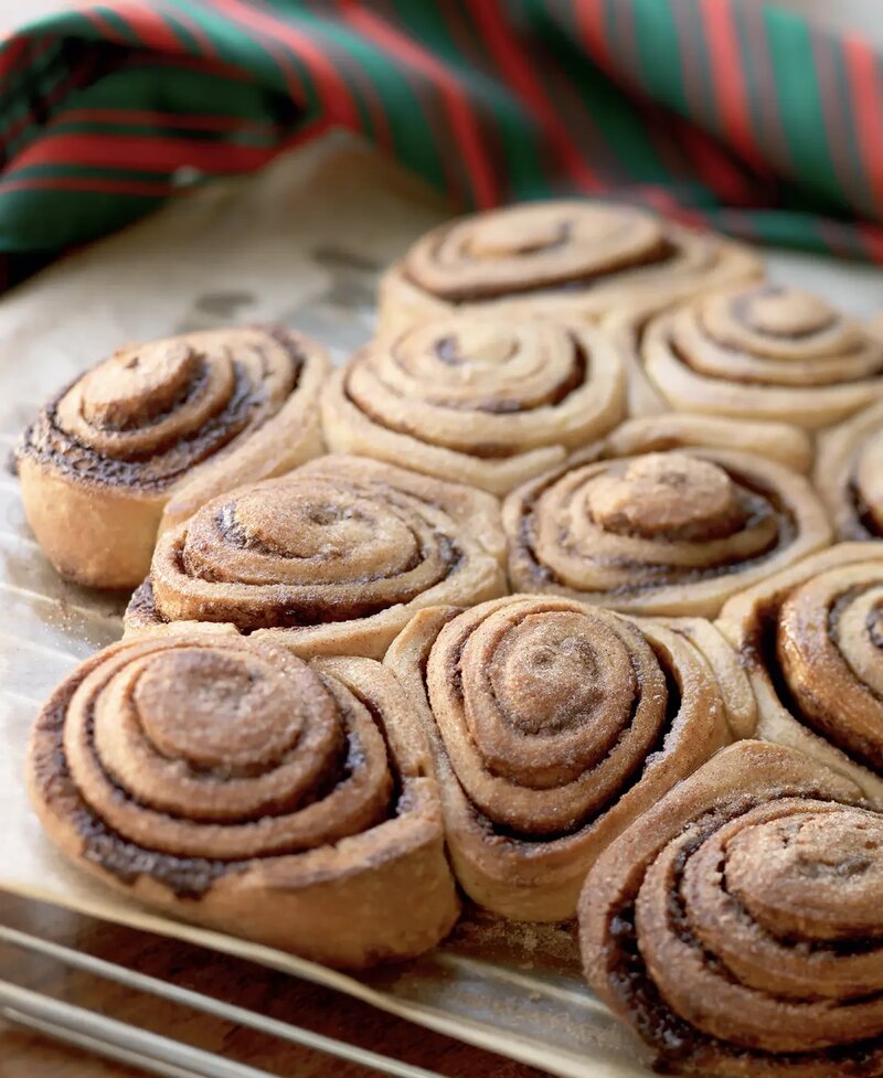 A close-up of freshly baked cinnamon swirls arranged in a glass baking dish, with a festive red and green striped cloth in the background, perfect for the 12 days of Christmas.