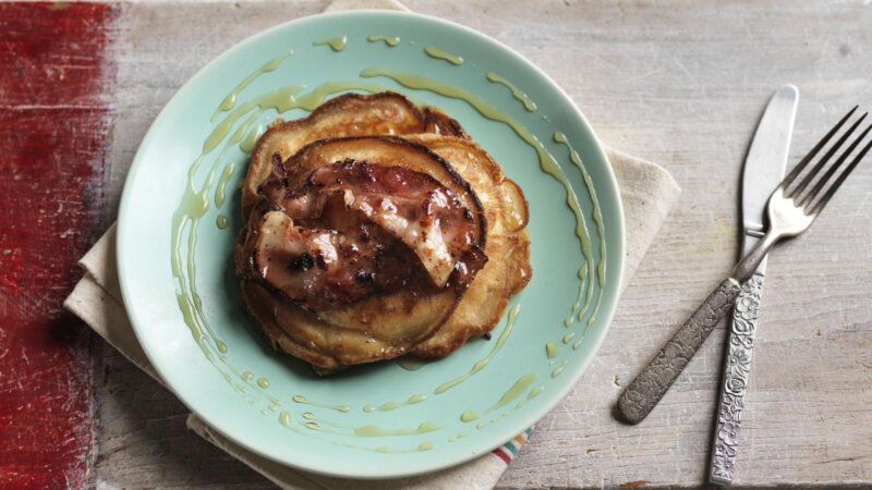 A stack of banana & bacon pancakes topped with grilled bacon and drizzled with syrup on a light green plate next to a fork and knife on a light wooden table, reminiscent of the Hairy Bikers' homey style.