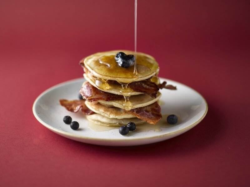 A stack of banana & bacon pancakes topped with grilled bacon and drizzled with syrup on a light green plate next to a fork and knife on a light wooden table, reminiscent of the Hairy Bikers' homey style.